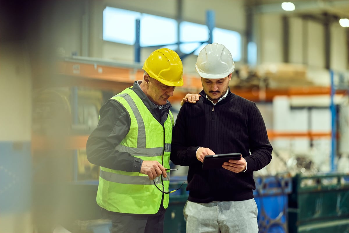 Two factory workers wearing hard hats and safety vest review production data on a tablet while collaborating on an inspection in an industrial warehouse