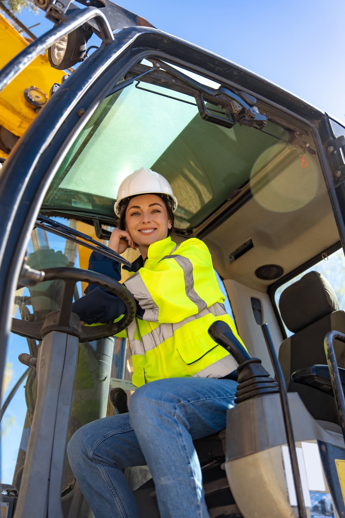 Cheerful female driver wearing white hardhat and safety vest sitting in excavator cabin and smiling